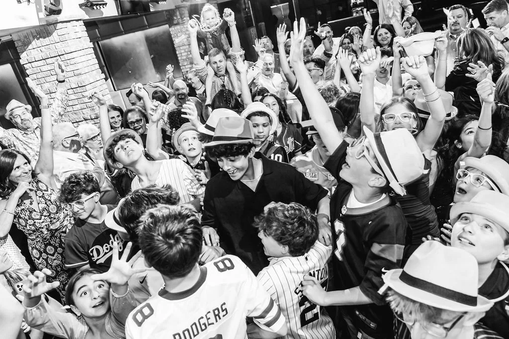 A large group of children and adults celebrating together at a party or event, many wearing hats, glasses, and sports jerseys, with some raising their hands and smiling.