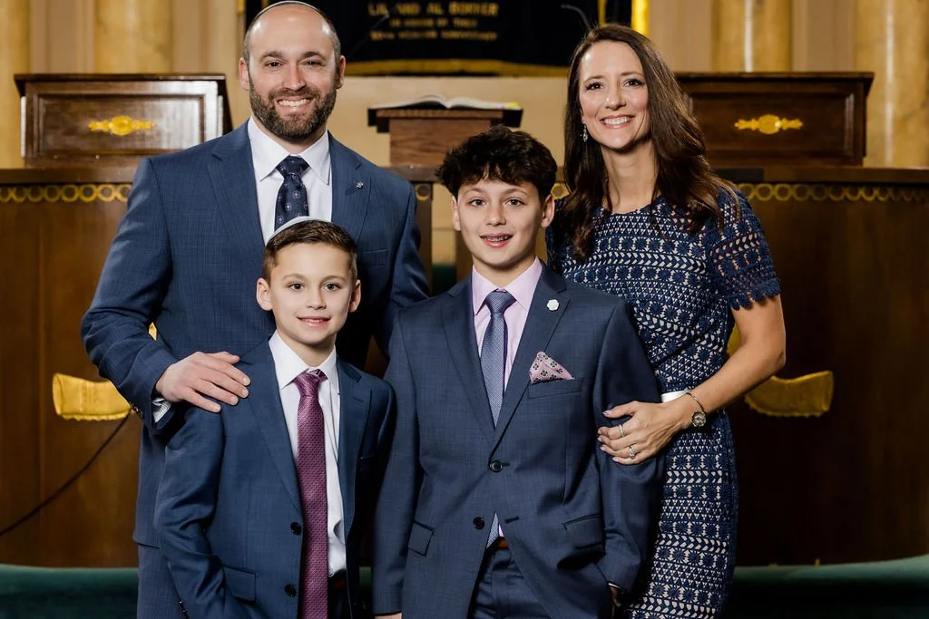 A family of four, two adults and two children, dressed in formal attire, posing together in a church or similar setting with wooden pulpit and gold accents behind them.