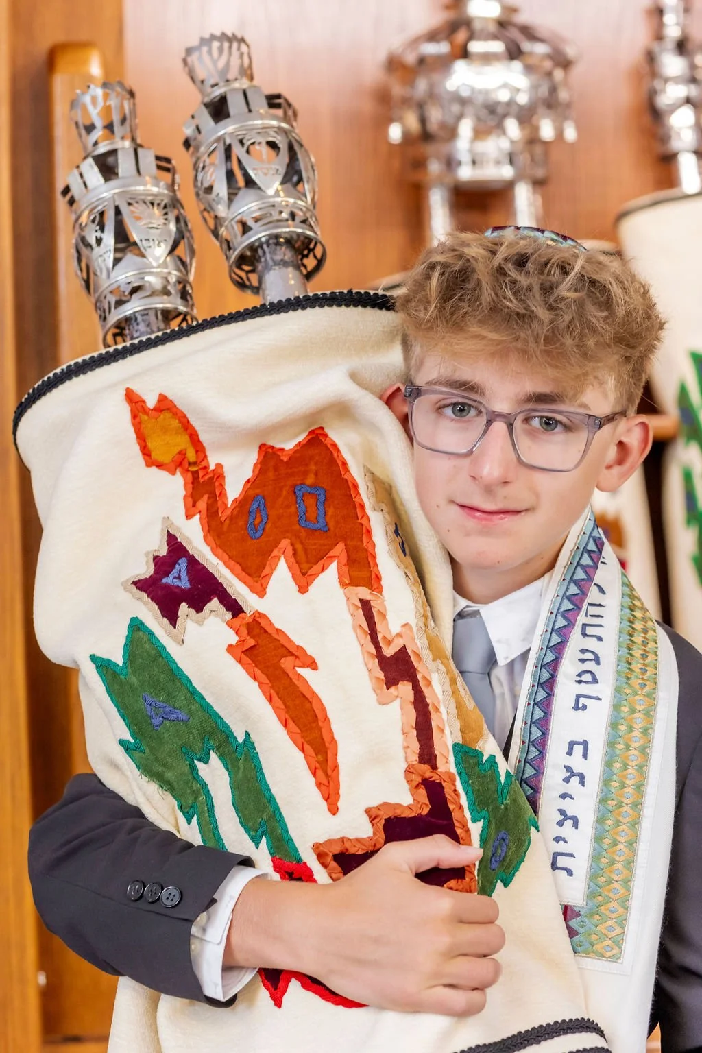 A young boy with curly hair, glasses, and a suit holds a tall Jewish Torah scroll decorated with embroidered images of a camel, a lightning bolt, and a star, and a colorful prayer shawl over his shoulder.