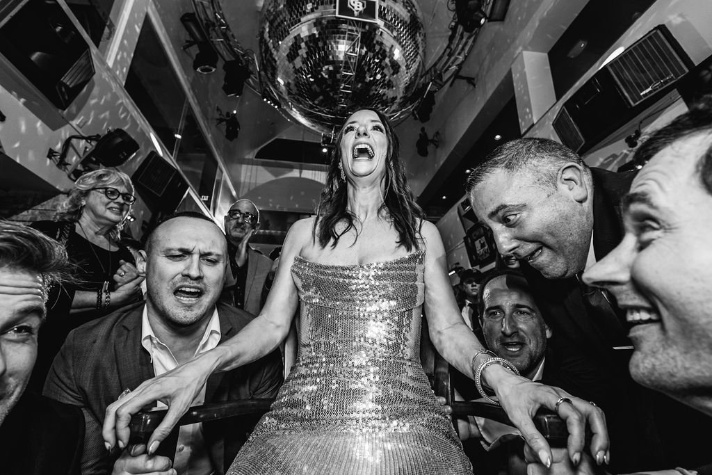 A woman in a shiny dress is laughing and surrounded by people at a lively party or celebration.