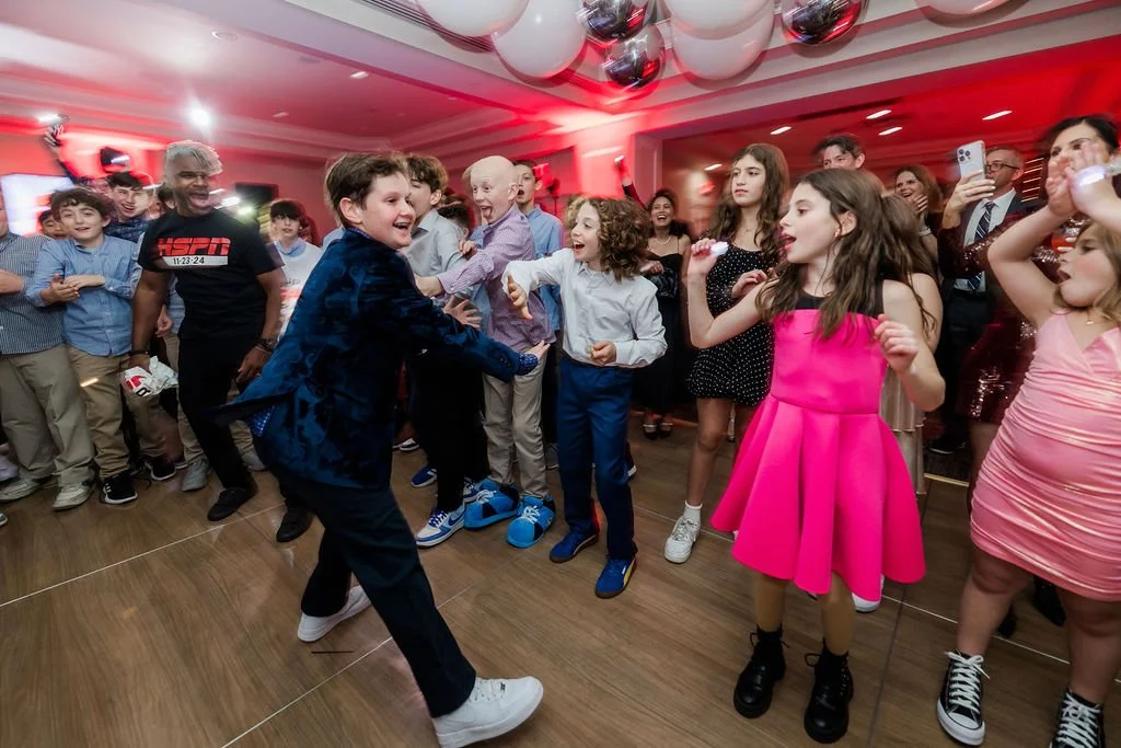 Kids dancing and having fun at a party or celebration, with some adults watching and taking photos.