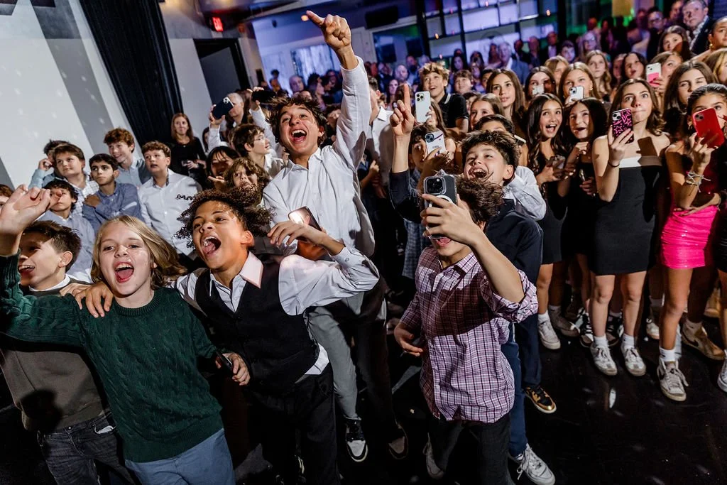 A large group of excited children at a school event, some taking photos and cheering.