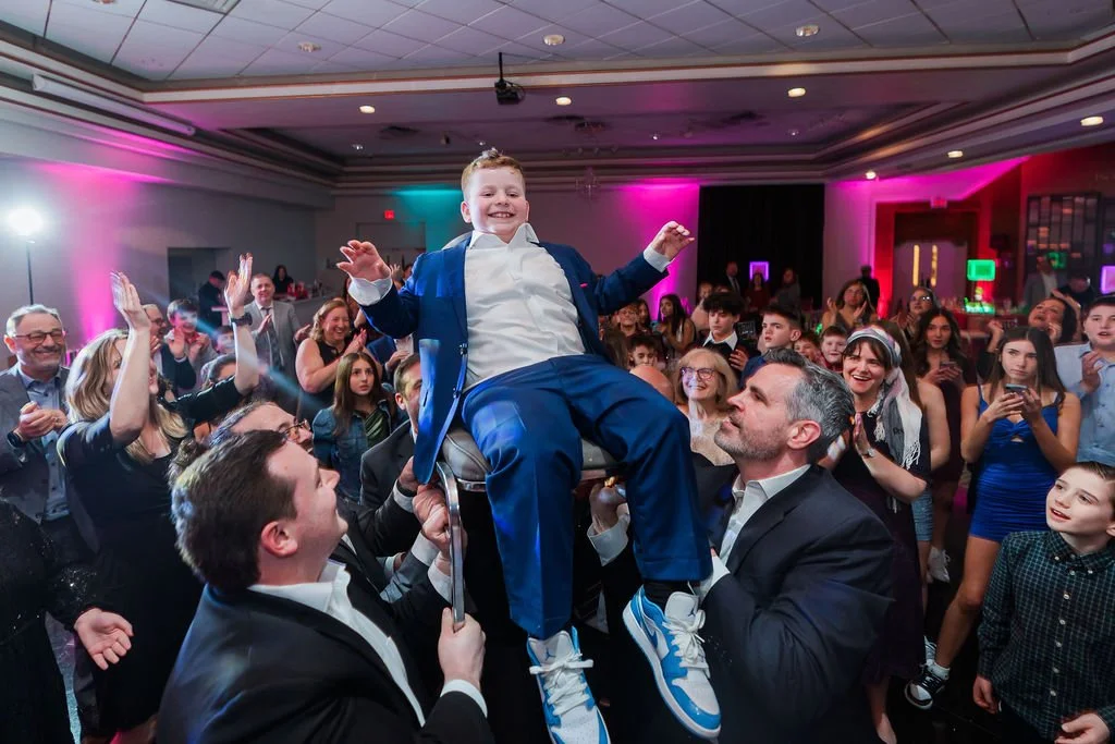A boy in a blue suit being lifted on a chair at a party, surrounded by smiling and clapping people in a decorated event hall with colorful lighting.