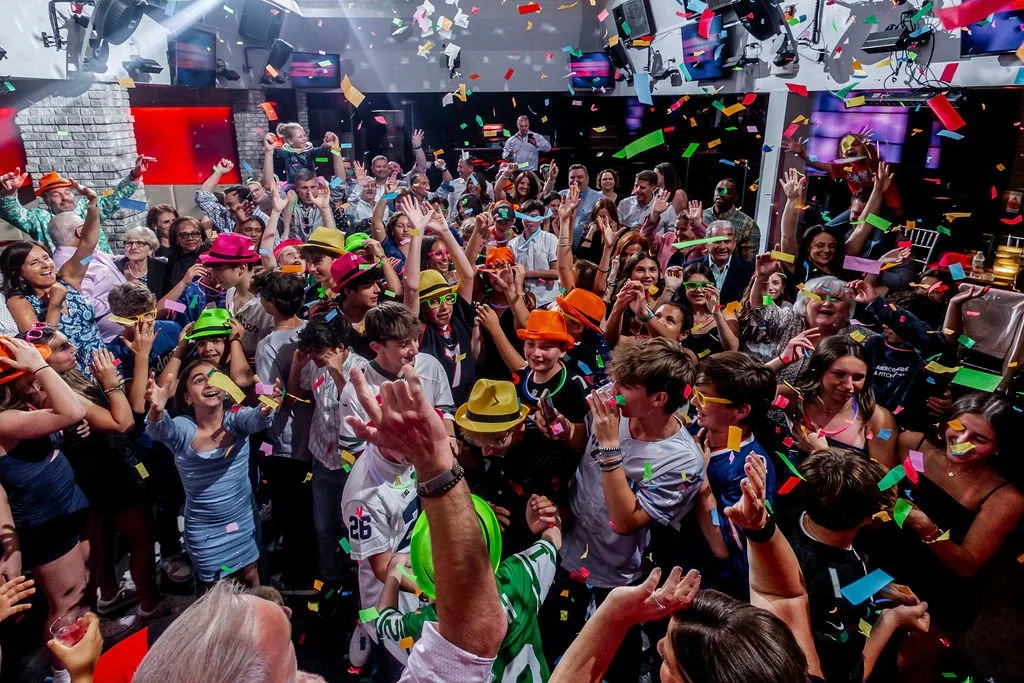 Children and adults celebrating at a party with confetti falling from above, some wearing colorful hats and glasses, in a lively indoor setting.