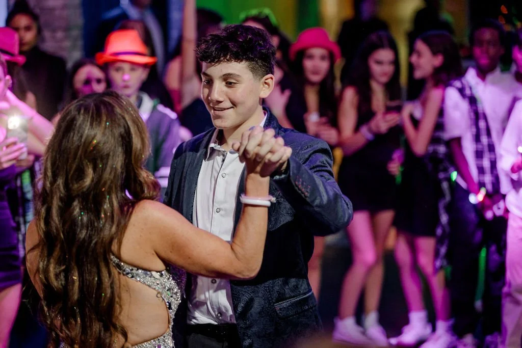 A young man and woman dance together at a party, surrounded by teenagers in colorful attire and party hats.