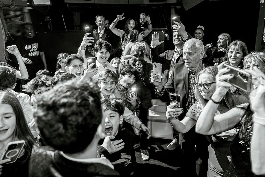 A lively group of people, including children and adults, gathered around a man in a suit who appears to be the focus of attention. Many are smiling, holding smartphones, and taking photos or videos, suggesting a celebratory or special event.