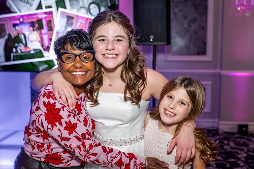 Three smiling women hugging each other at an indoor party with purple lighting and photographs on a wall in the background.