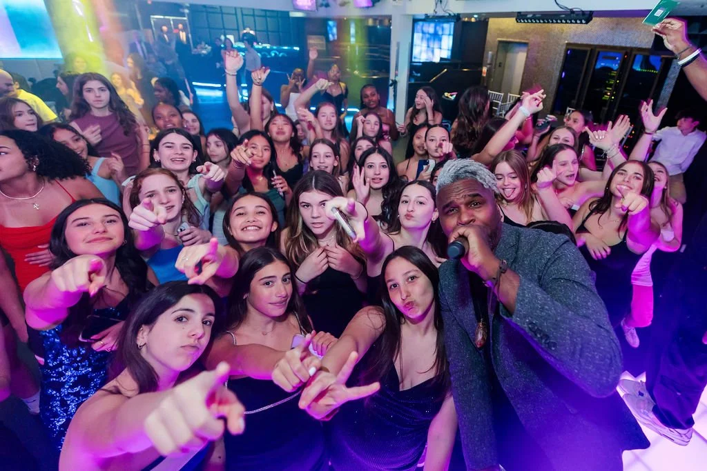 A large group of young girls and a man with gray hair holding a microphone gather at a lively party or dance event with colorful lighting.