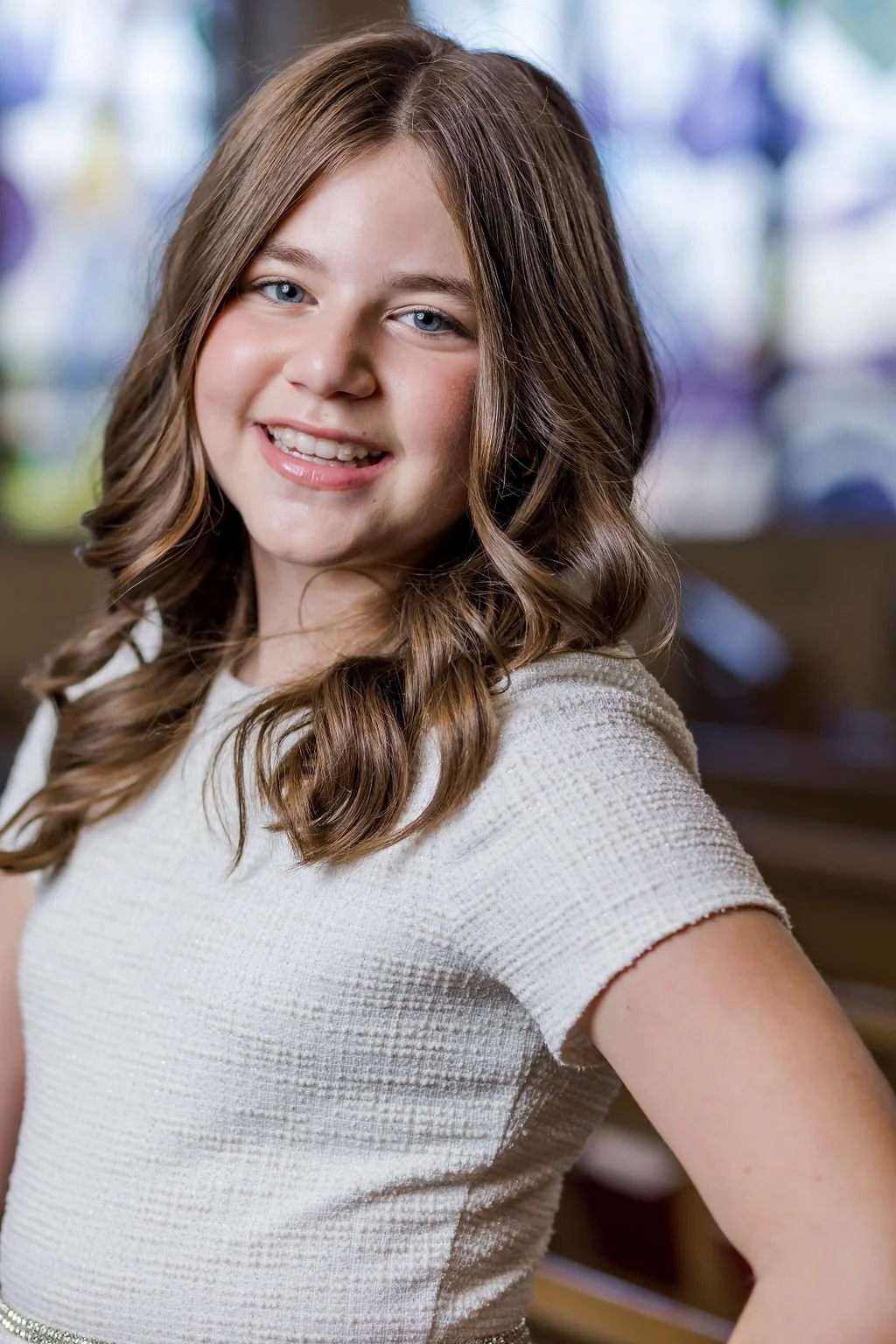 A young girl with wavy brown hair and blue eyes smiling at the camera, wearing a cream-colored textured top, indoors with a blurred background.
