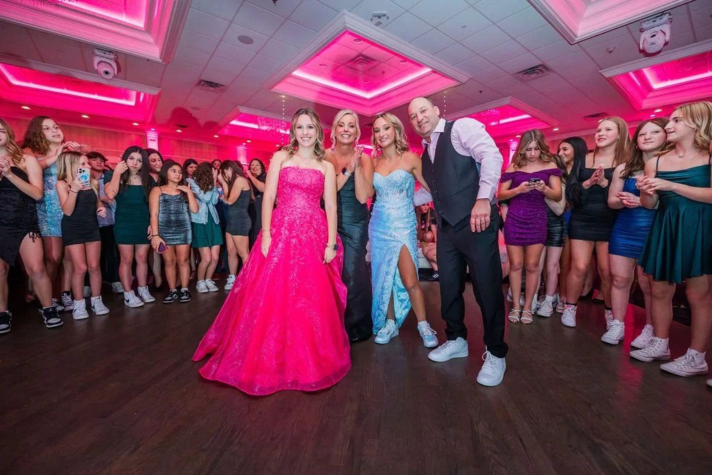 Group of young women and two adults at a dance party in a decorated ballroom, pink ceiling lights, girls in formal dresses, some taking photos and dancing.