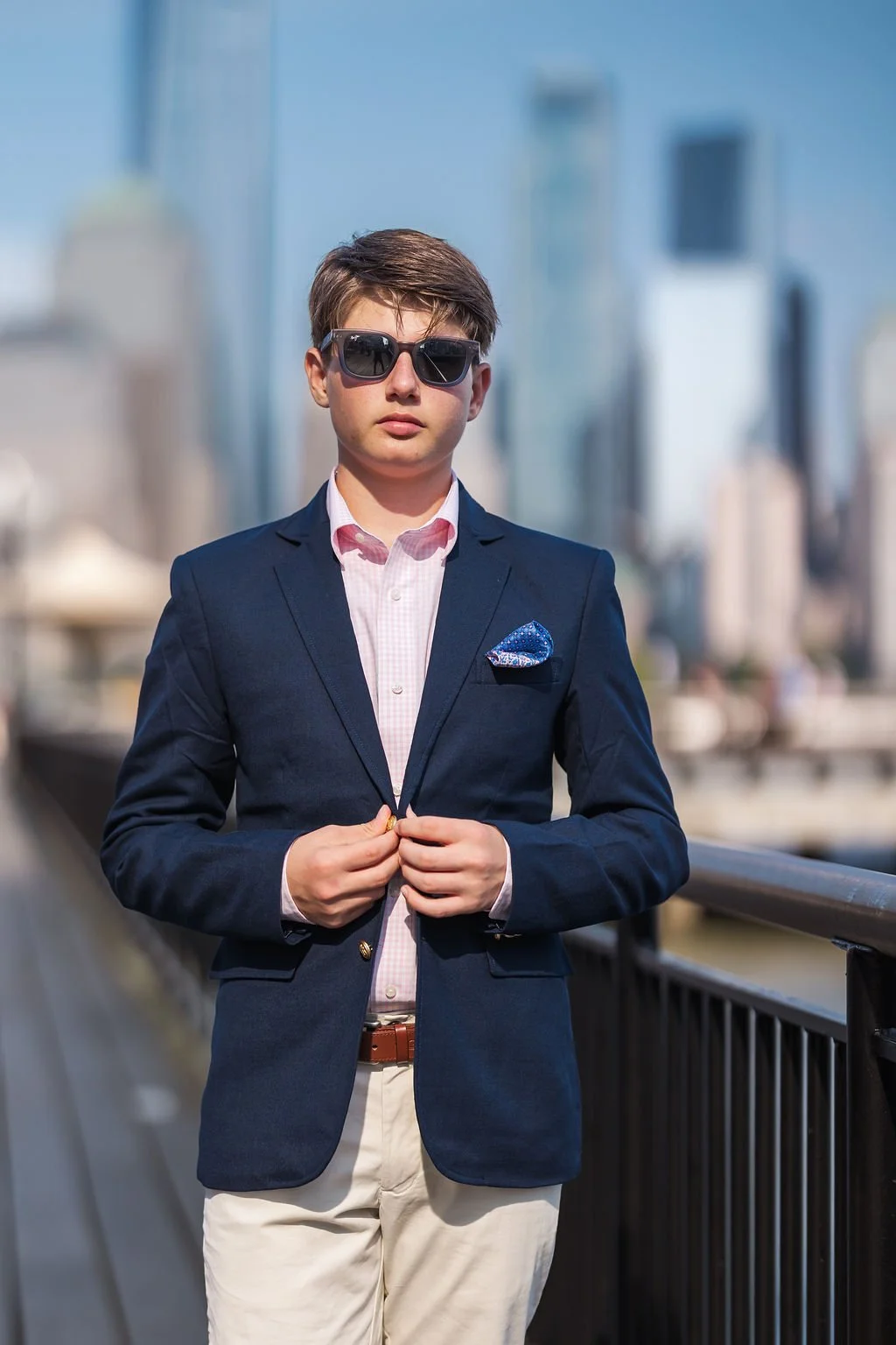 A young man in a navy blazer, light pink shirt, beige pants, wearing sunglasses, standing outdoors on a city street with modern buildings in the background.