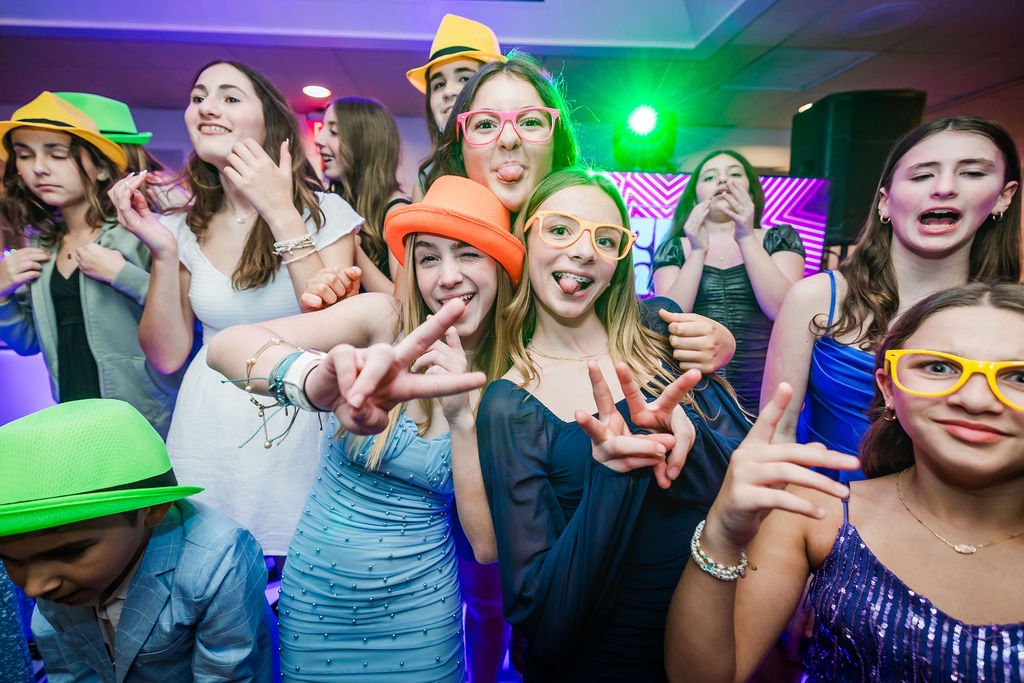 Group of young girls at a party, some wearing colorful hats and glasses, making playful gestures, with colorful lighting and a decorated background.