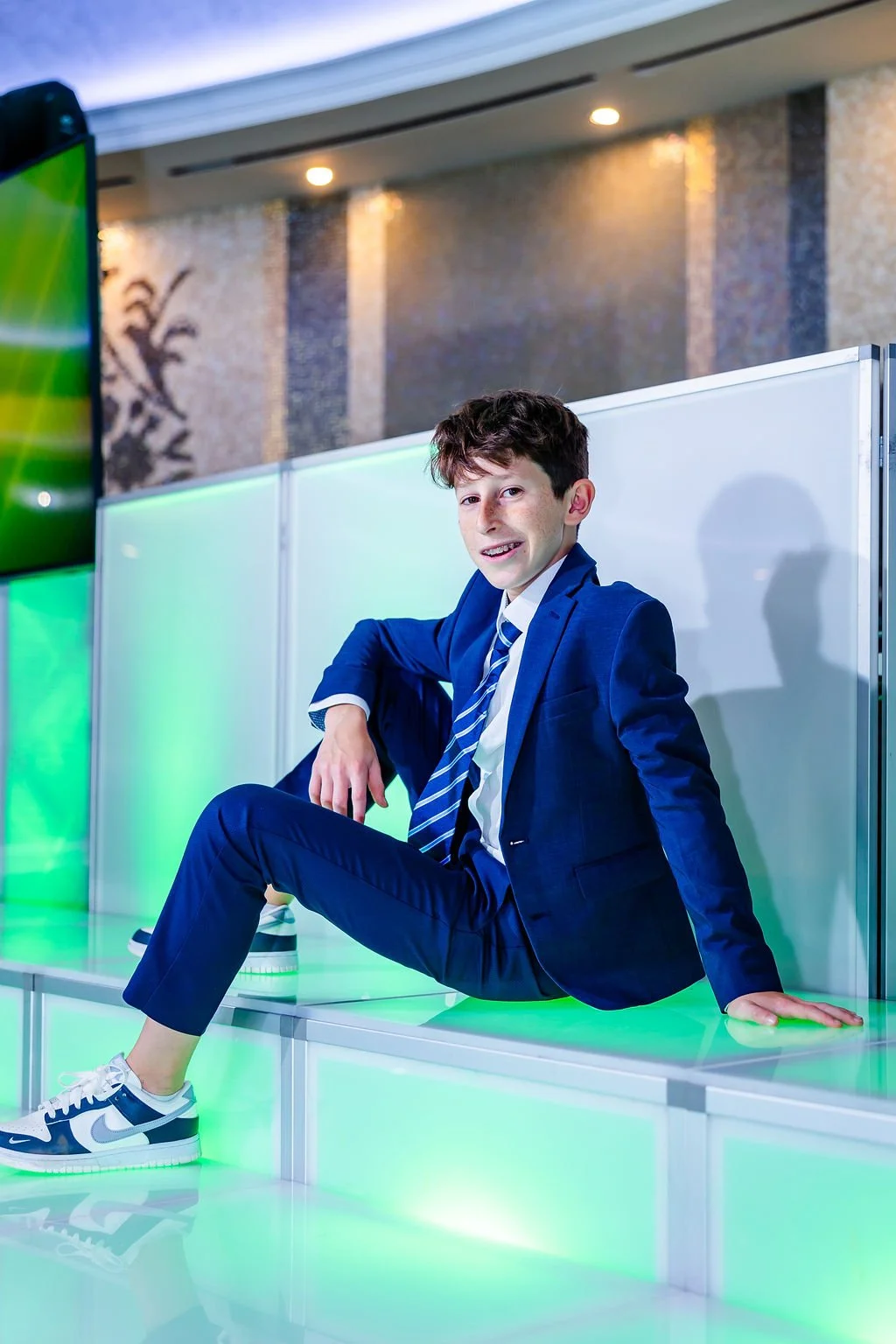 A young boy in a blue suit and striped tie sitting on a illuminated green bench with a white partition behind him at an indoor event.