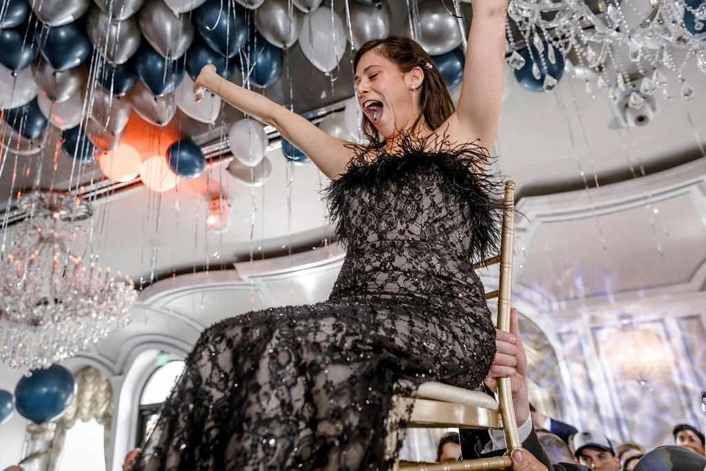 Woman in a black lace dress celebrating on a chair at party decorated with balloons and chandeliers.