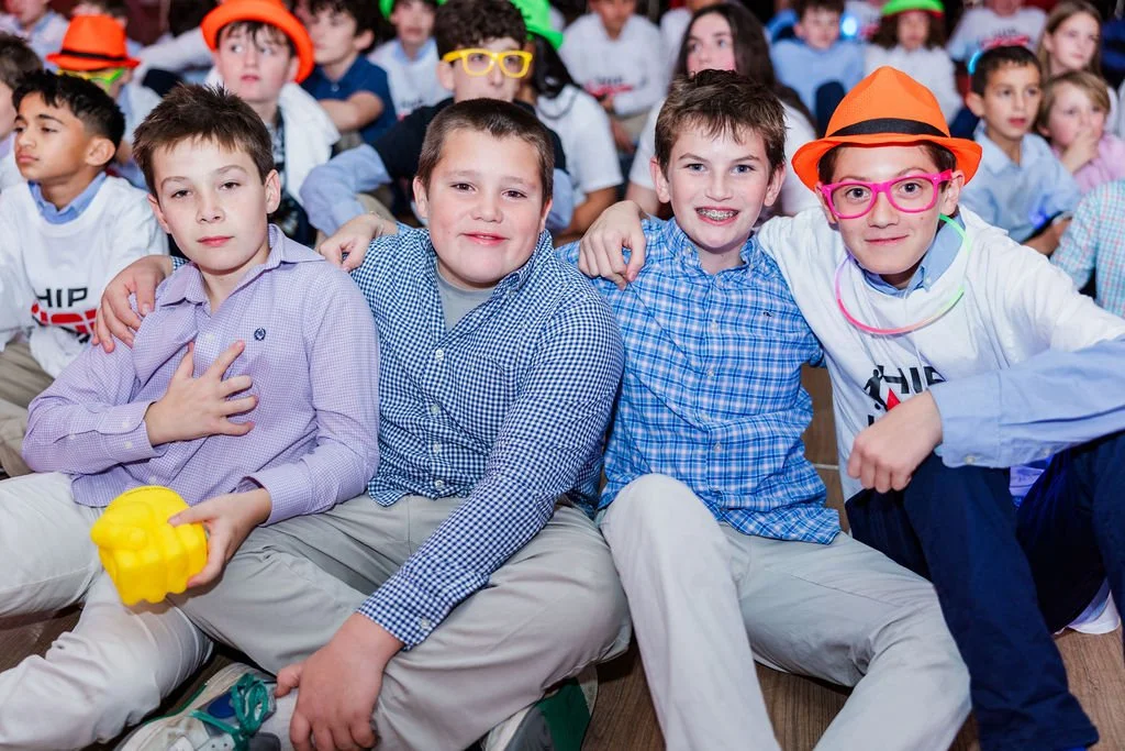 Four young boys sitting on the floor in a crowded room, smiling at the camera. They are dressed in casual clothes, with one wearing a bright orange hat and pink glasses, and another holding a yellow plastic toy.