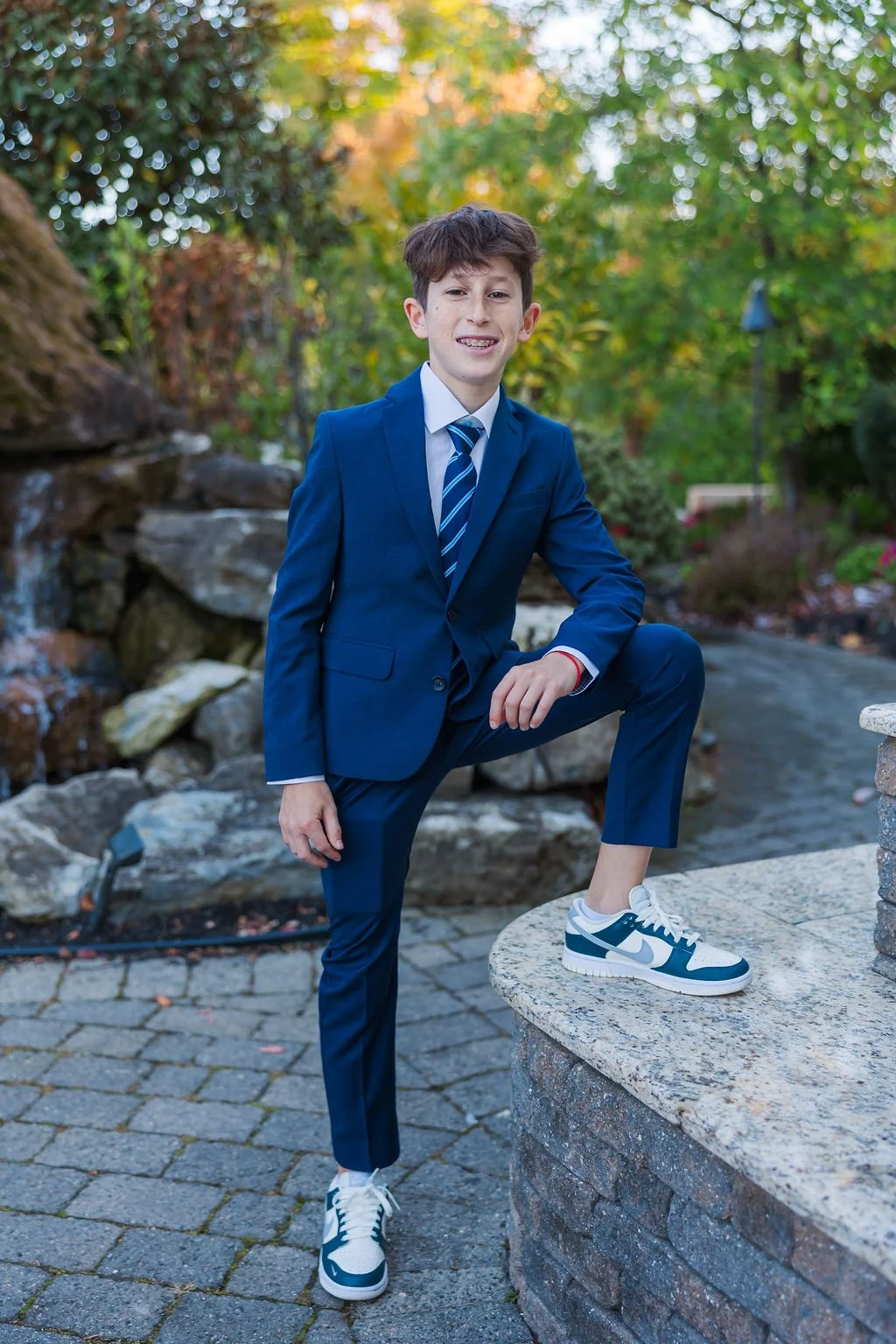 A young boy in a blue suit, tie, and white sneakers with blue accents, posing outdoors on a stone feature with a natural background of trees and rocks.