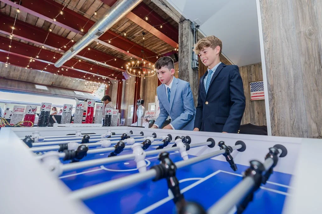 Two boys in suits playing foosball at an indoor arcade or entertainment venue decorated with string lights and American flags.