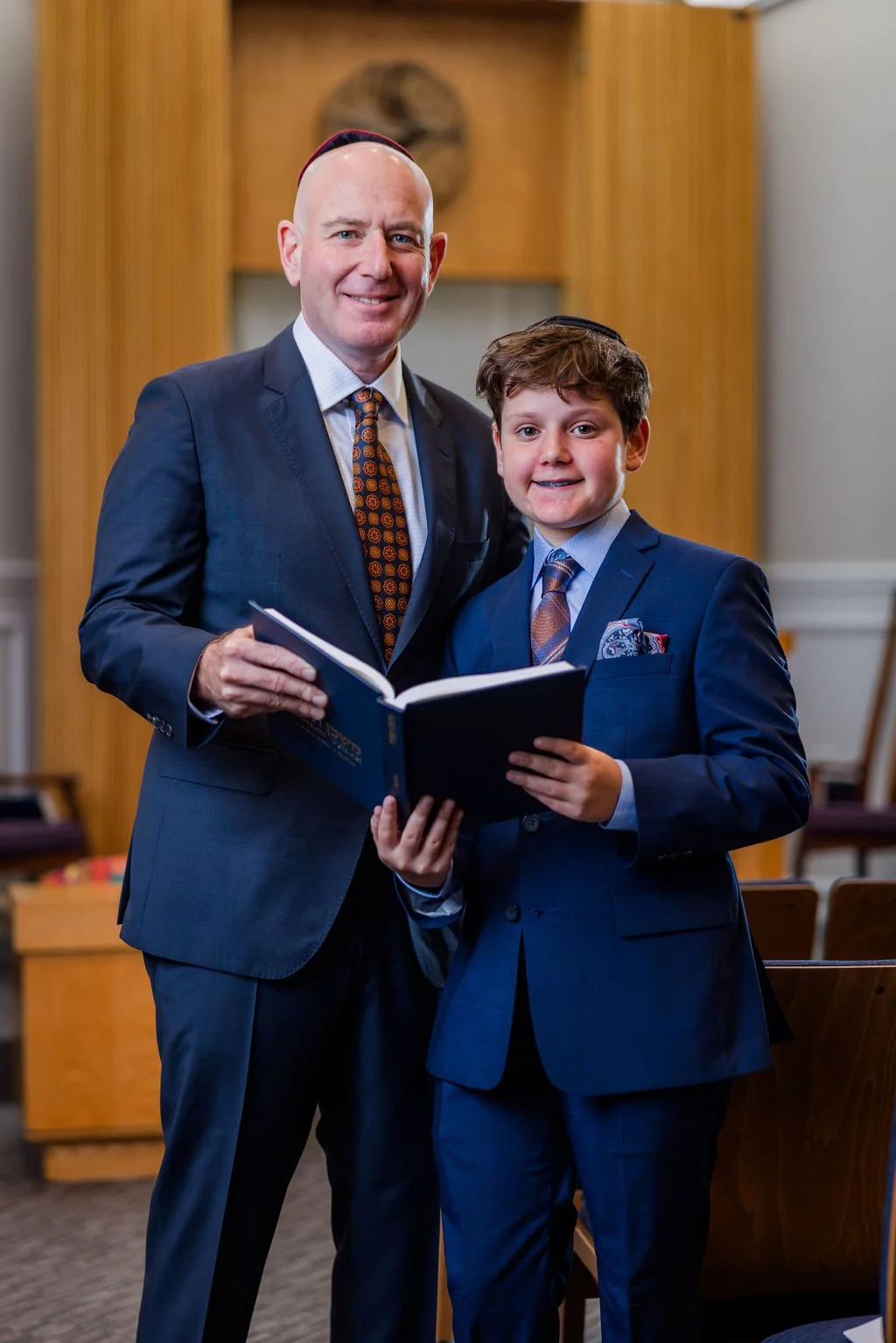 A man and a boy dressed in suits standing indoors, smiling, and holding a book together.
