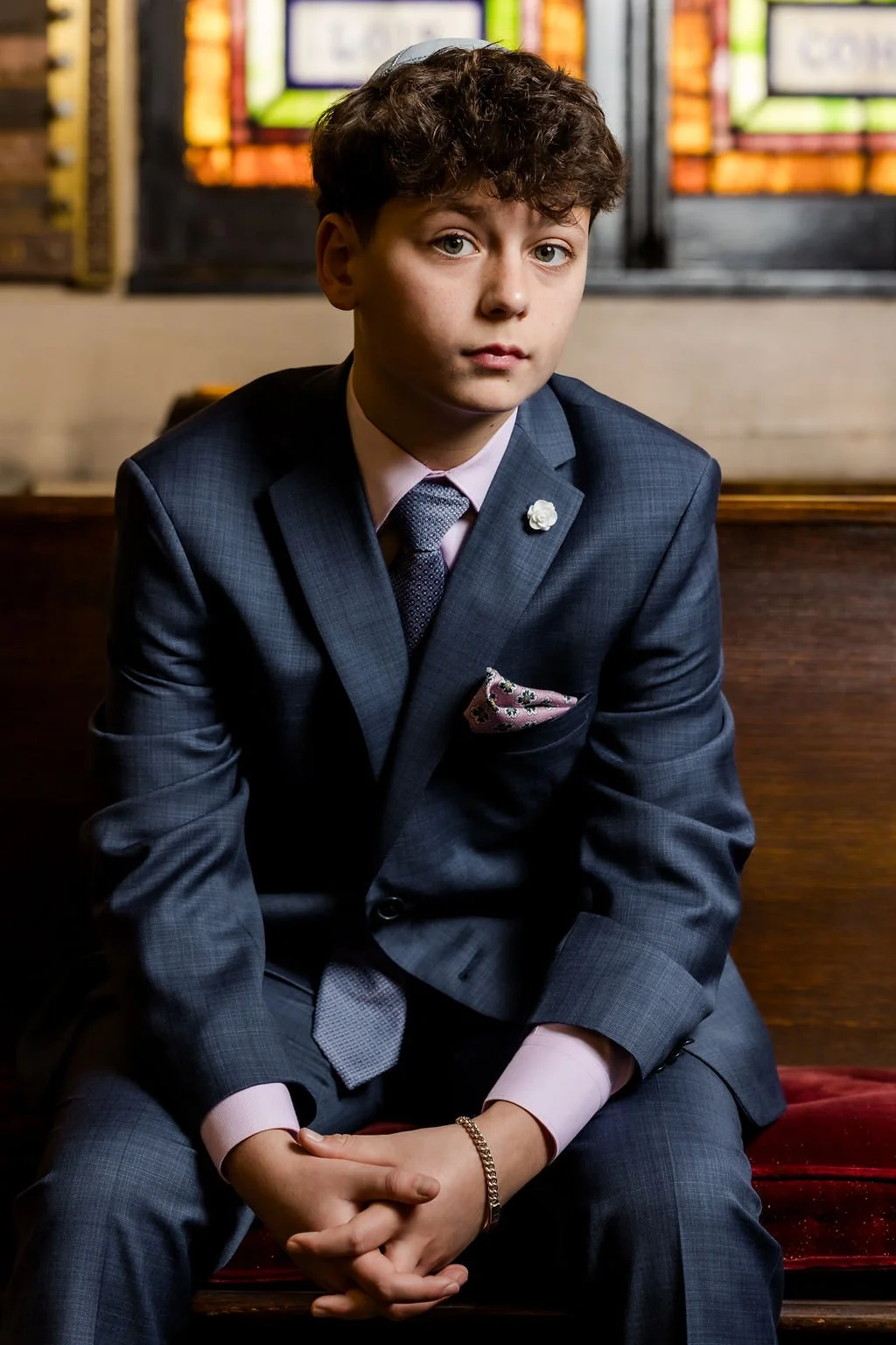 A young boy with curly hair sitting on a wooden bench, wearing a dark blue suit with a pink shirt, a patterned tie, a pink pocket square, and a gold bracelet, in a room with stained glass windows in the background.