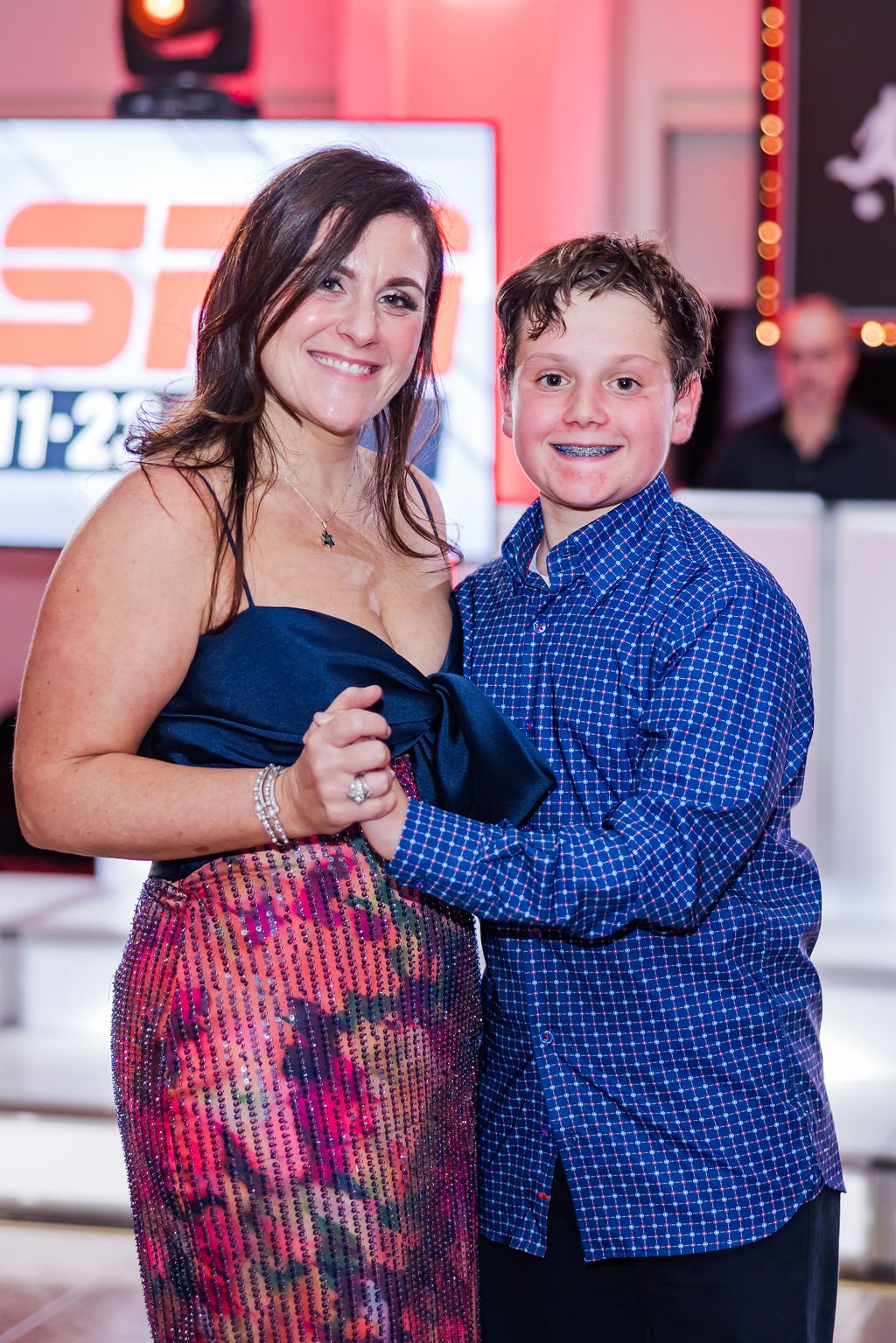 A woman and a young boy smiling and dancing together at an event, with the woman wearing a blue dress and the boy in a blue checkered shirt, holding hands.