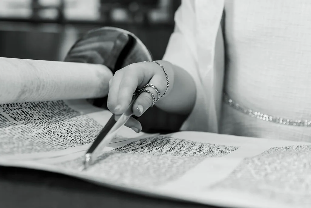 Person reading a large open book or manuscript, with a pen in hand, dressed in a light-colored top and wearing rings and a bracelet, in black and white.