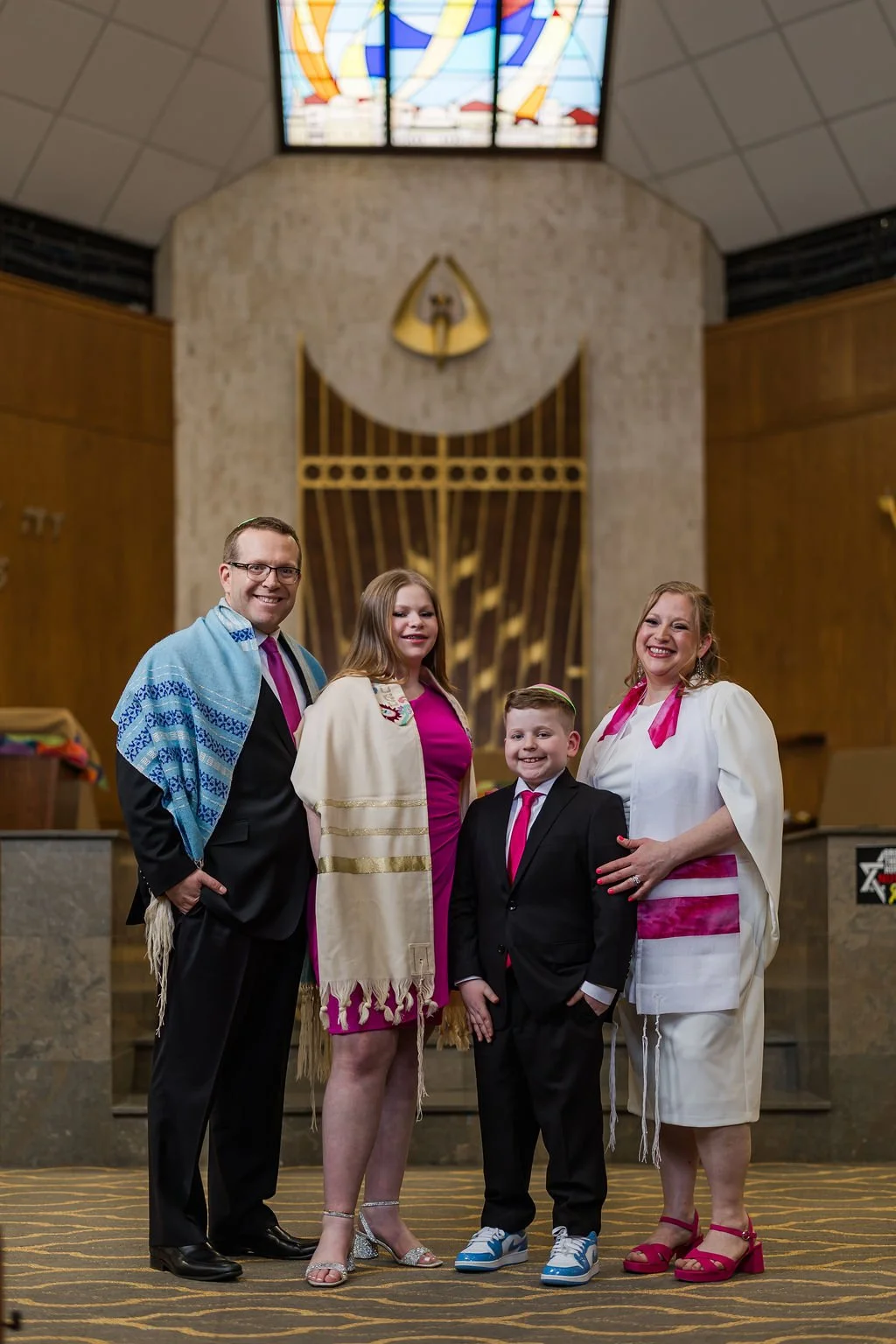 A family of four standing inside a temple, dressed in formal attire with religious shawls over their shoulders, smiling for a group photo.