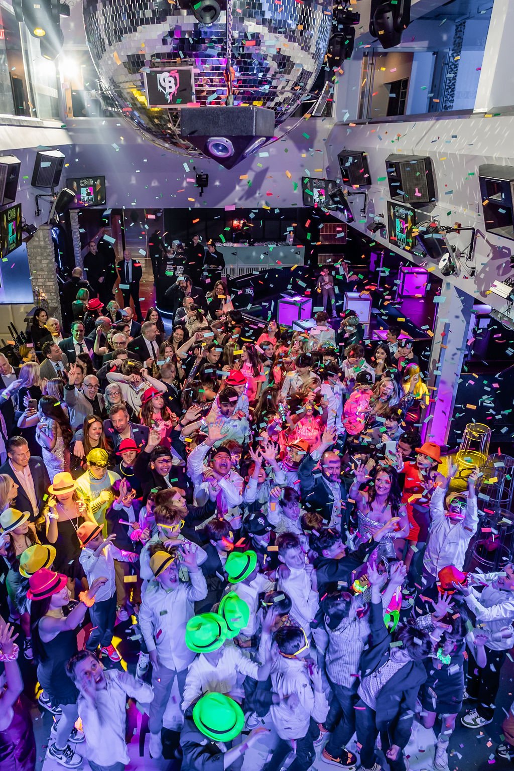 People dancing and celebrating at a party with colorful lighting, confetti, and a disco ball ceiling.