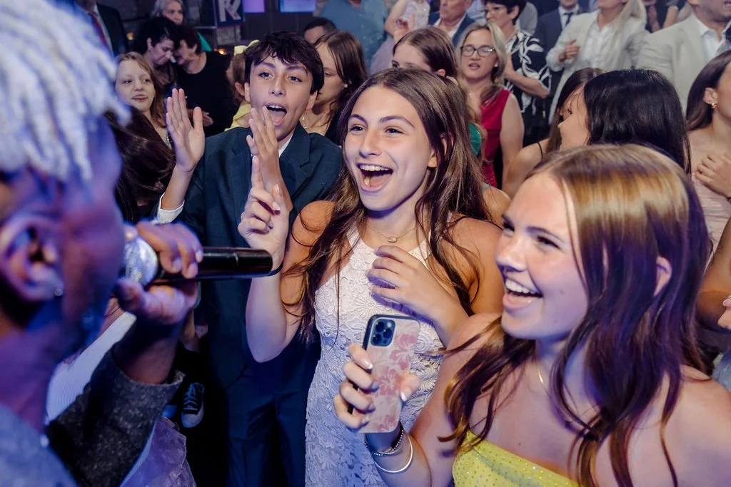 A group of young people at a lively event, smiling and laughing while singing or talking to a person holding a microphone, with many more attendees in the background.