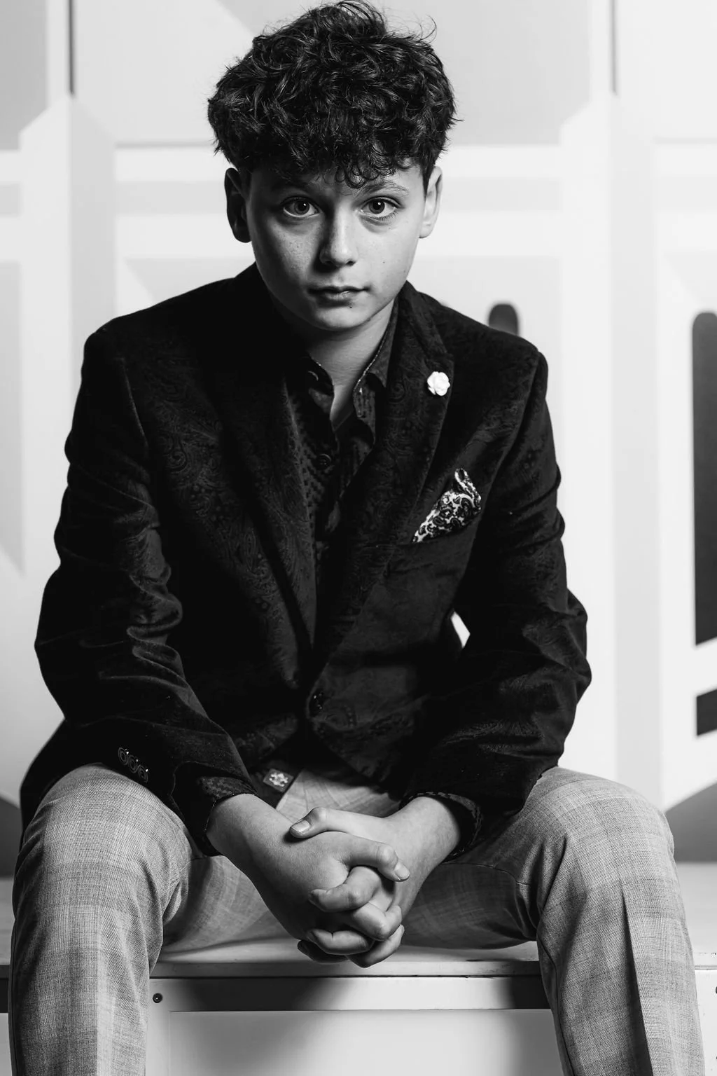 A young man with curly hair, wearing a dark blazer with a patterned pocket square, sitting with his hands clasped in front of him, against a background of white paneling, in black and white.