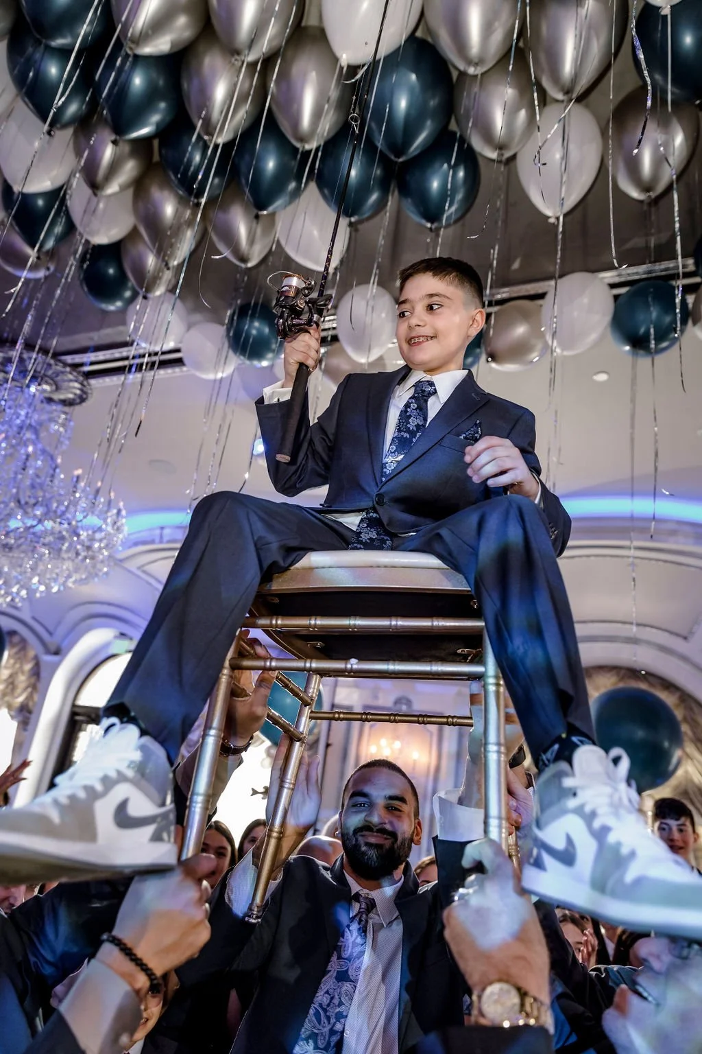 A young boy in a suit sitting on a chair held up by adults at a celebration or party with balloons overhead.