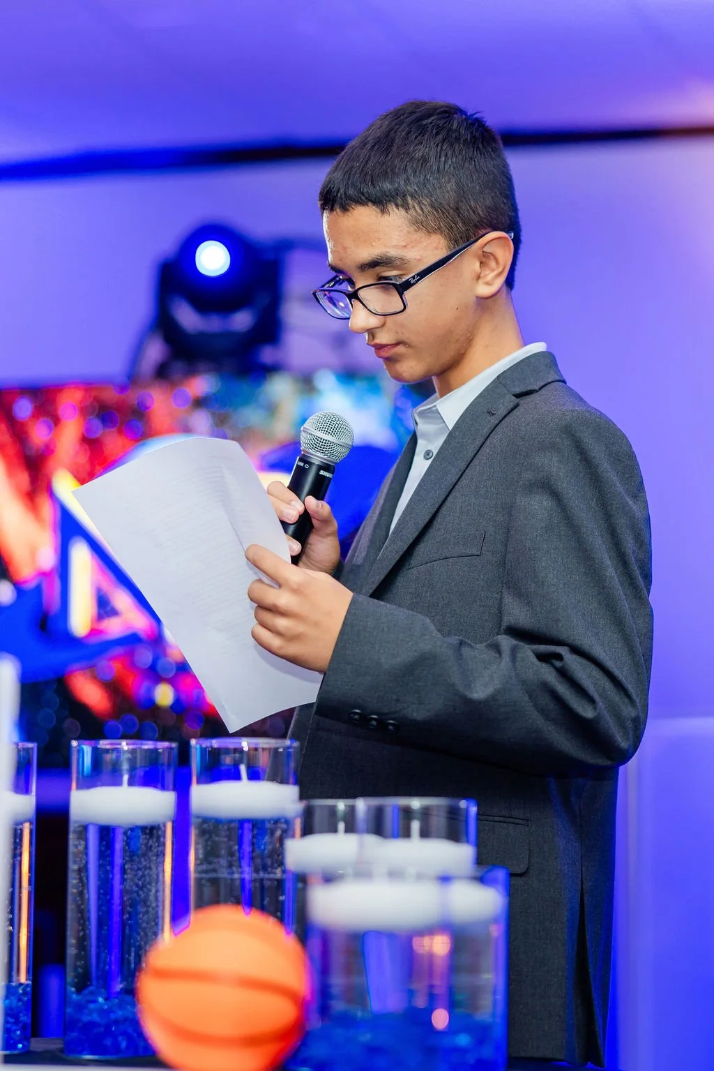 A young man in a gray suit and glasses reading from a paper into a microphone at a formal event, with multiple glasses with candles and an orange ball in the foreground, and colorful lighting in the background.