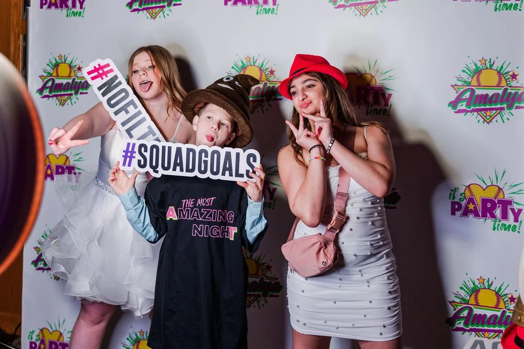 Three children posing for a photo at a party, holding signs that say '#GUILTY' and '#SQUADGOALS.' They are in front of a backdrop with colorful party-themed graphics and text.