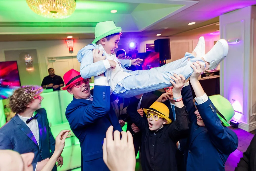 Children dressed in colorful suits, hats, and glasses partying and lifting a young boy in a suit and hat in the air at a festive event with colorful lighting.