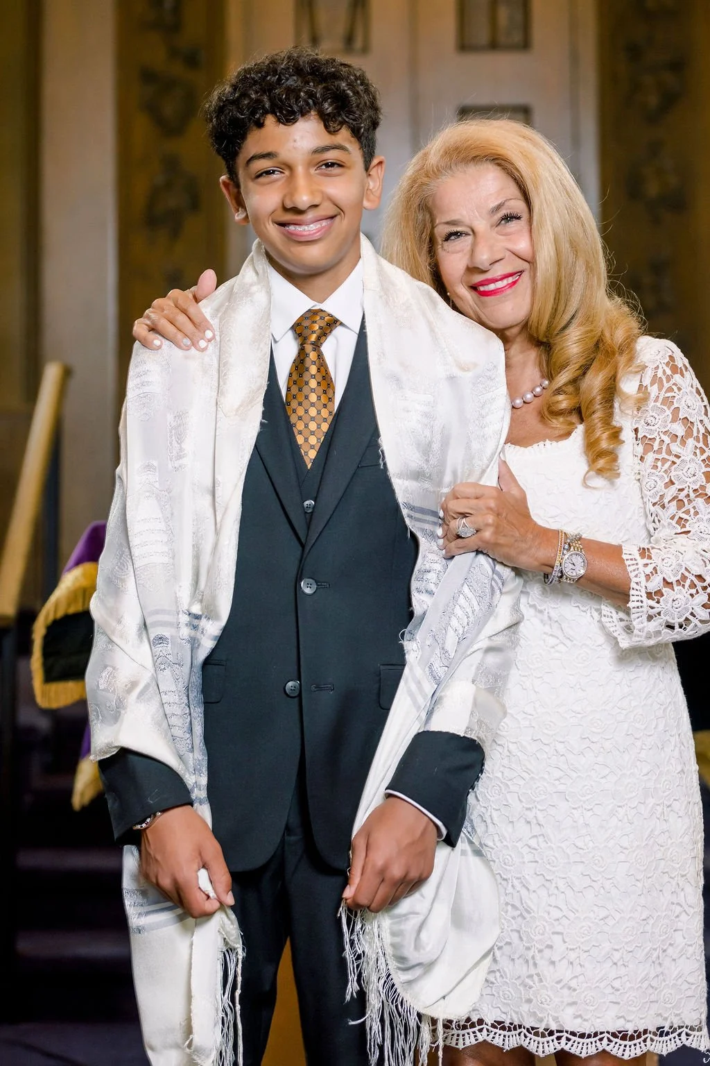 A young man in a dark suit with a patterned tie and a white shawl, smiling, standing next to an older woman with long blonde hair in a white lace dress, also smiling, in an indoor setting.