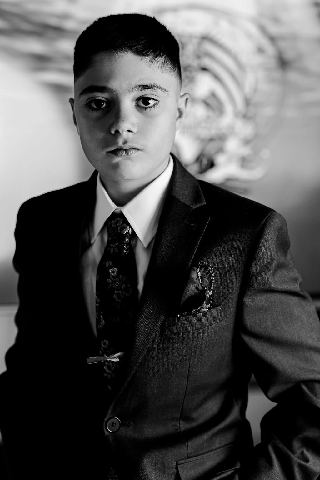 Black and white portrait of a young boy with short hair, wearing a formal suit, tie, and pocket square, looking directly at the camera with a serious expression.