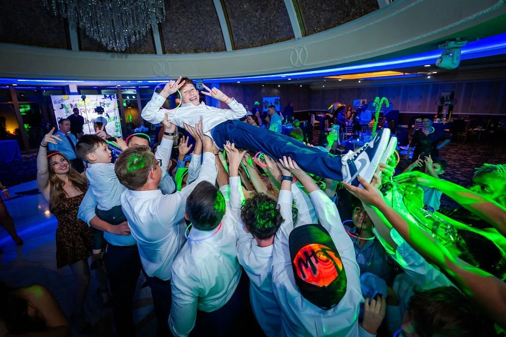 People at a party lifting a young man in the air while dancing, with colorful lighting and decorations in the background.