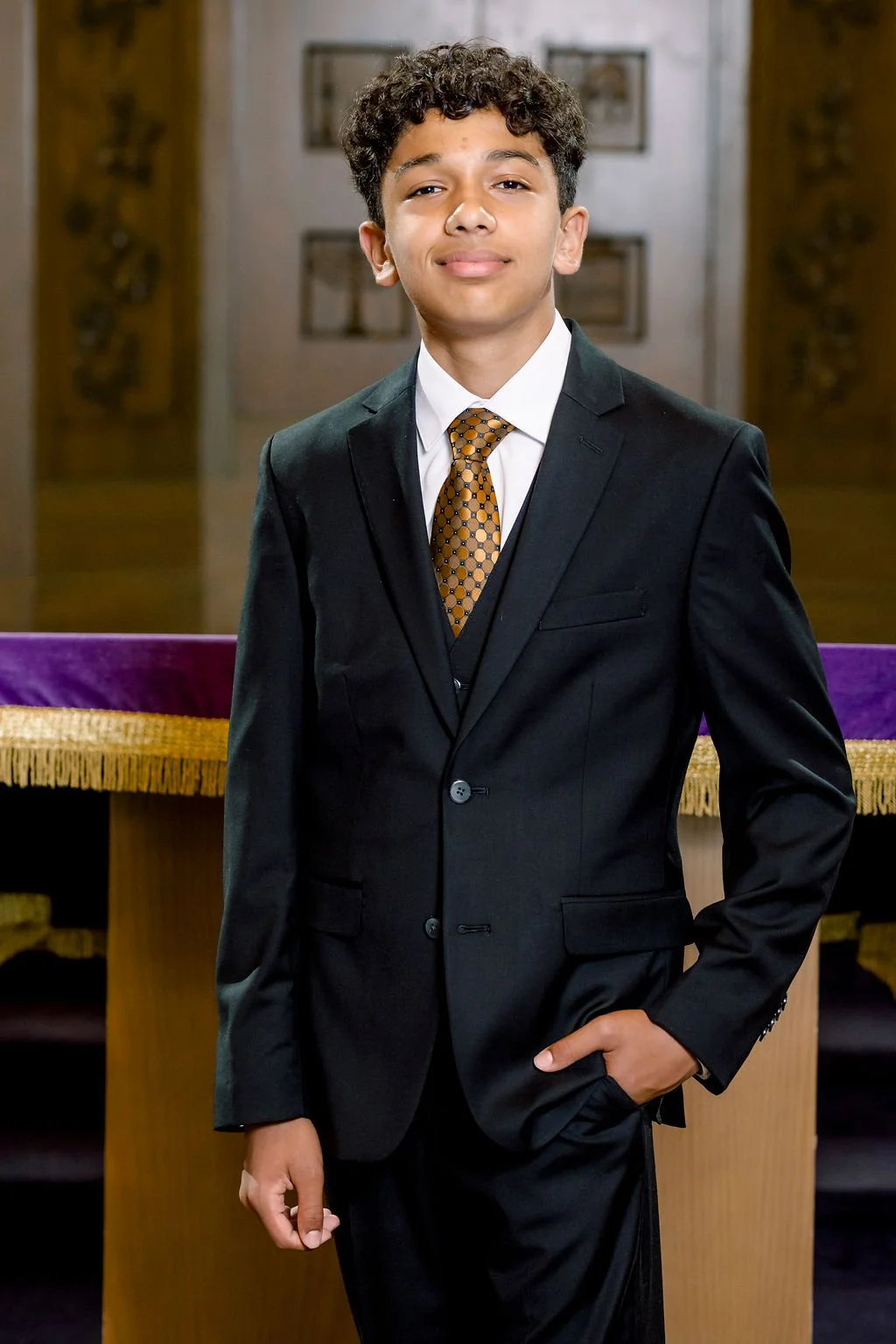 Young man with curly hair dressed in a black suit, white shirt, and a patterned tie standing in a formal setting.