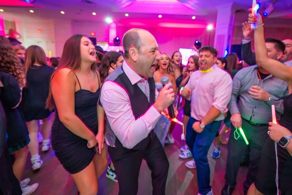 People dancing and singing at a lively party with colorful lights, some holding glow sticks, and a man with a microphone in the center.