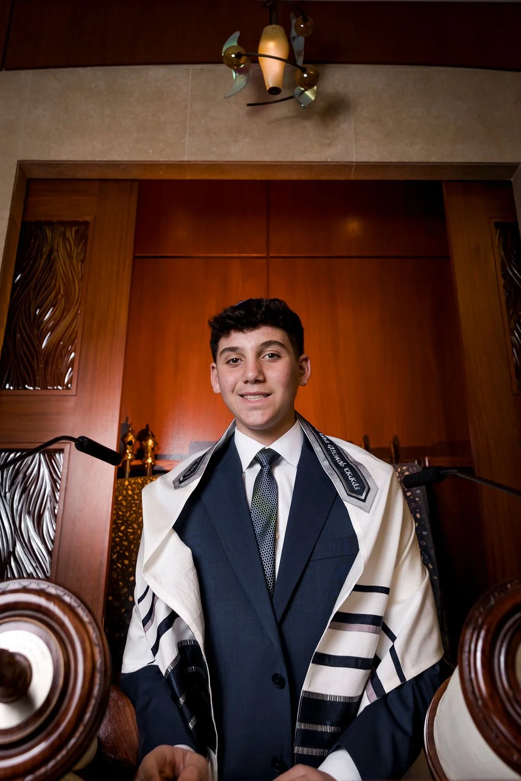Young man in religious or ceremonial attire standing inside a wooden room with ornate woodwork, smiling at the camera.