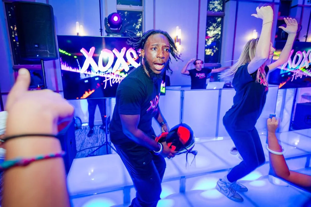 People dancing at a nightclub or party with colorful lighting, a DJ, and a large screen displaying 'XOXO' in the background.