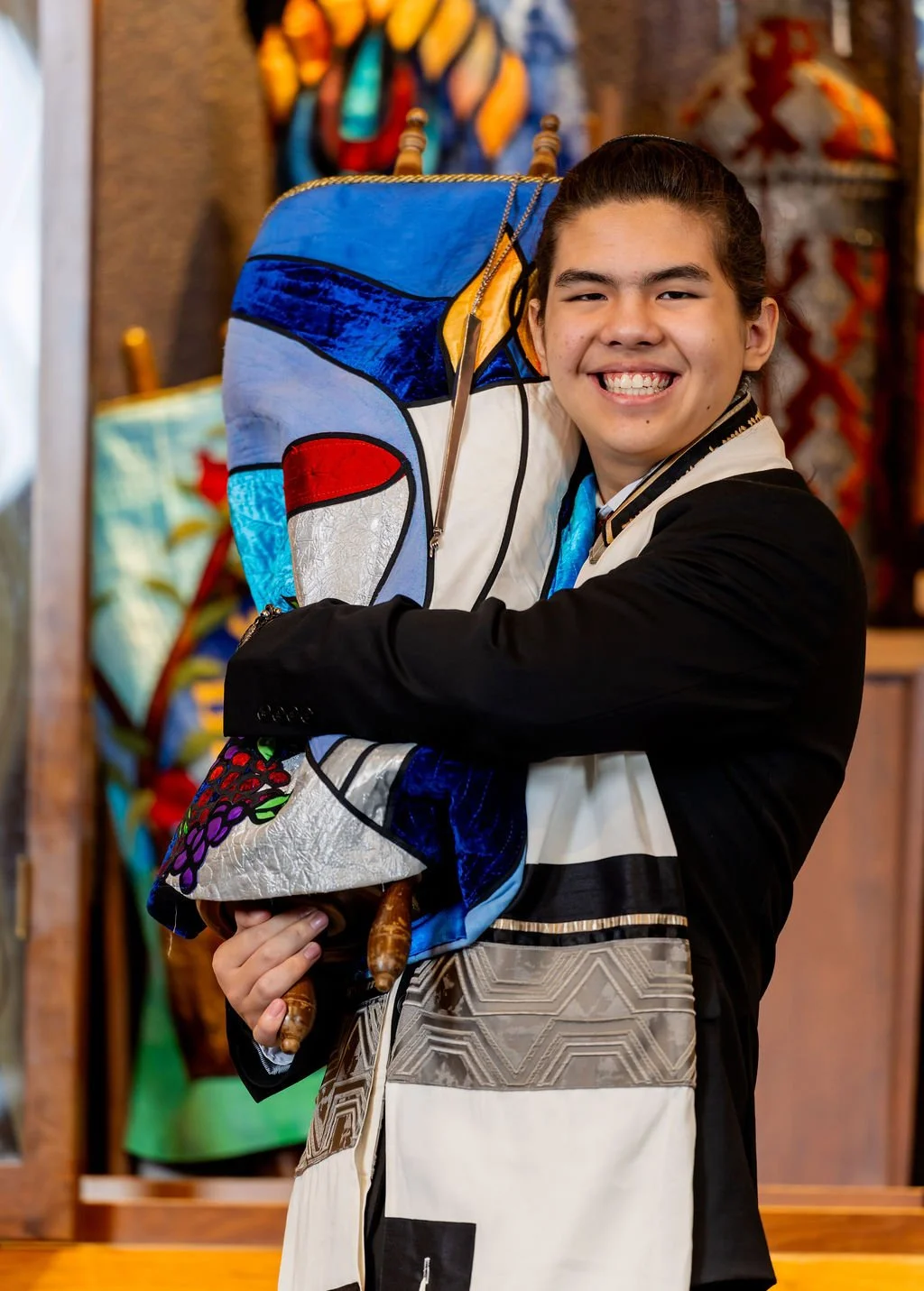 Young person smiling and proudly holding a colorful, decorated abalone shell in a shop with vibrant textiles and crafts in the background.