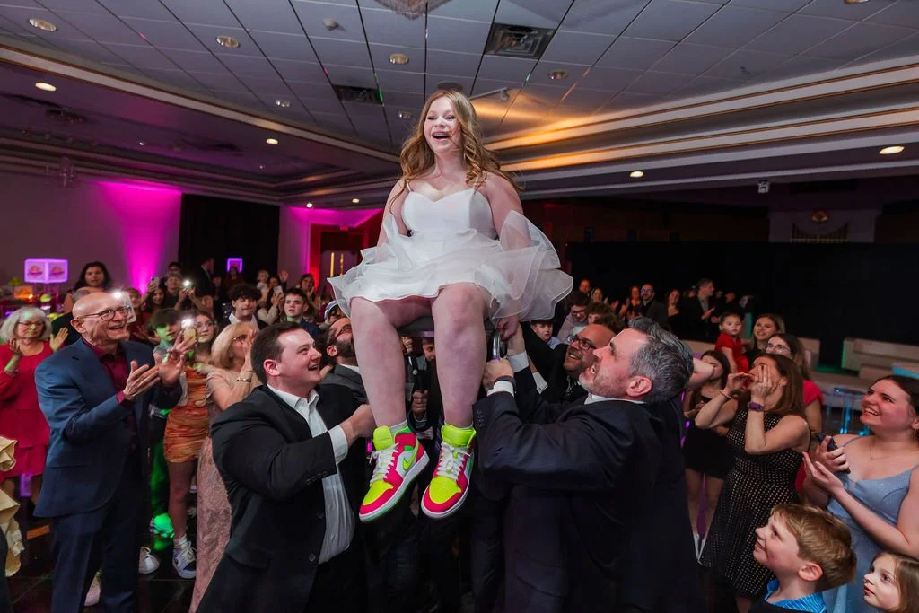 A young girl in a white dress being lifted by adults at a party with many people clapping and watching.