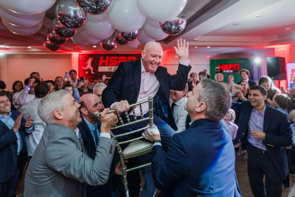 A group of people at a celebration, lifting a man in a chair while everyone laughs and cheers. The man in the chair is smiling and waving. The room is decorated with black, white, and silver balloons, and there are ESPN logos in the background.