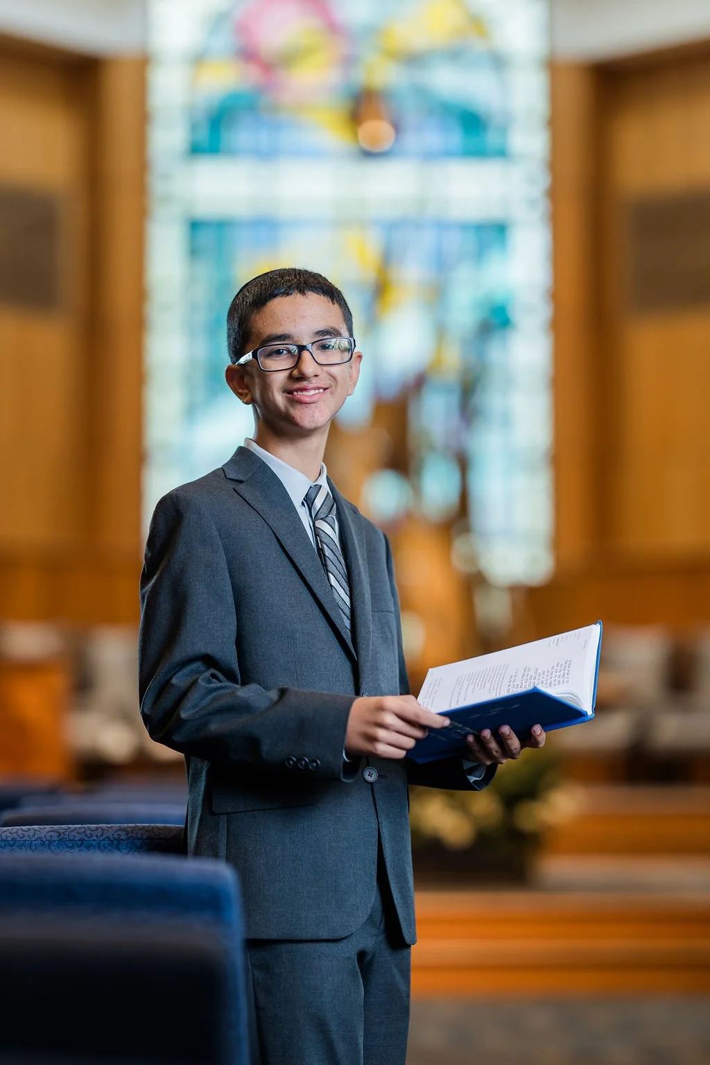 A young man in a gray suit and glasses standing inside a church, holding an open book and smiling at the camera.