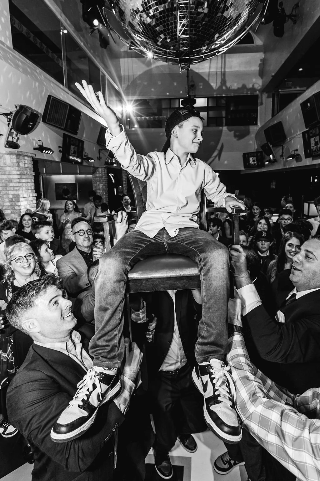 A young boy wearing a baseball cap and casual clothes is seated on a large chair being lifted by two men in suits at a lively party or celebration. The boy is making a peace sign with his right hand and smiling with his eyes closed. The crowd around 