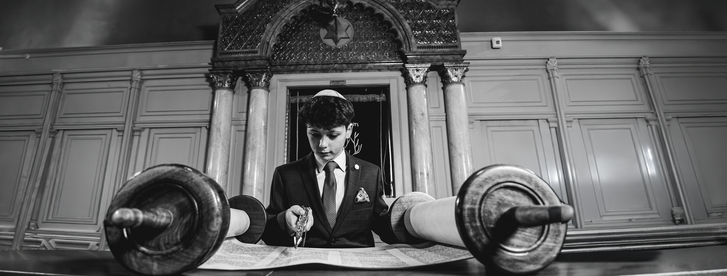 A young boy wearing a kippah and a suit, standing at a Torah scroll in a synagogue, with the Torah opened in front of him.