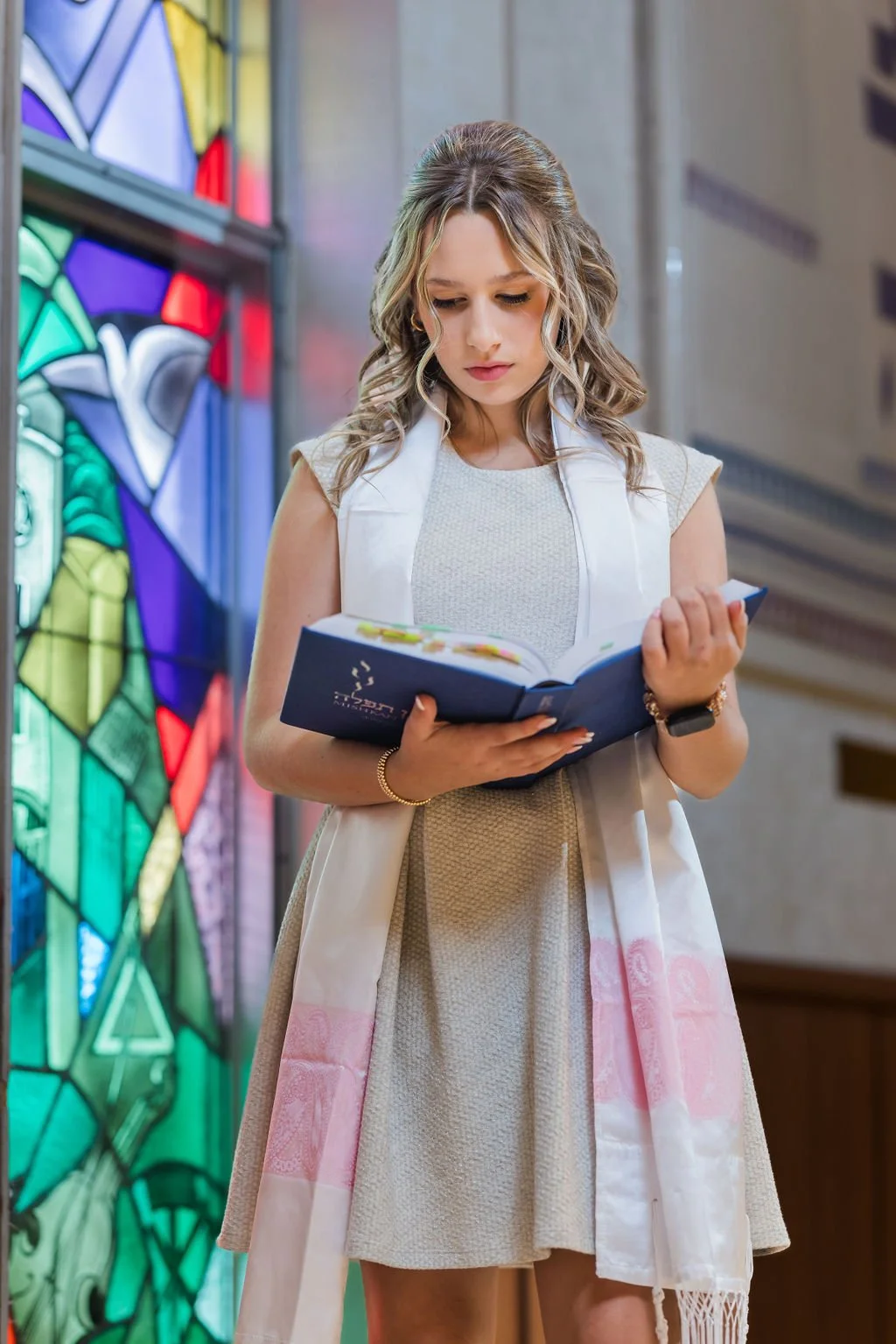 A young woman with wavy blonde hair reading a book inside a church or chapel near a colorful stained glass window.
