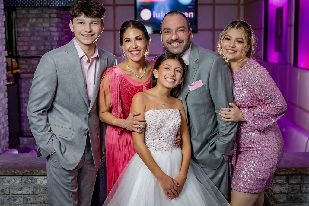 Family celebrating wedding reception, standing together, smiling in formal attire, bride in wedding dress, groom in suit, three women and one young man, colorful lighting in background.