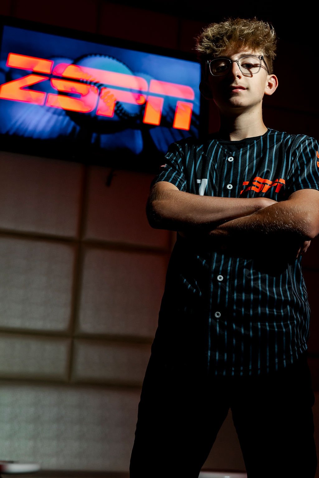 A young man with glasses and curly hair standing with crossed arms in front of a large screen displaying the word "ZOTI" in neon lights.