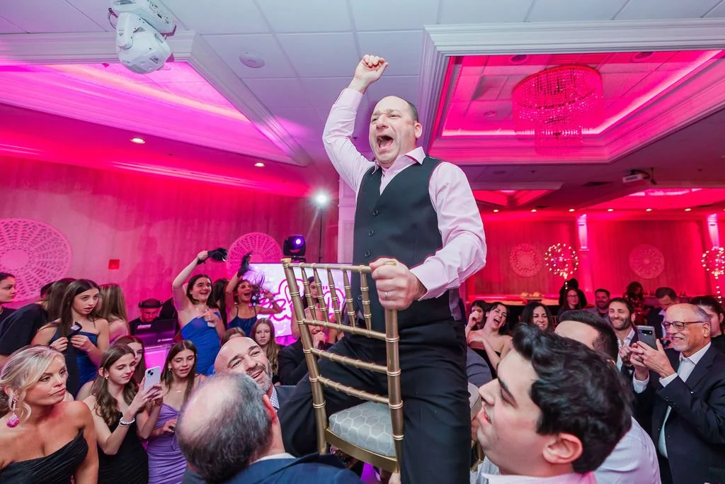 A man standing on a chair, holding one arm in the air, celebrating at a lively party with many people. The room is decorated with pink and purple lighting and elegant decor.